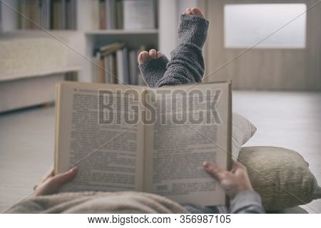 Woman reading a book with feet lying on pilow at home. Quarantine and isolation concept