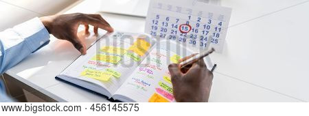 Businessman's Hand Checking Schedule In Diary With Calendar On White Desk