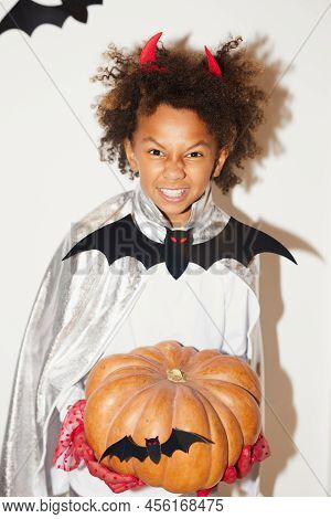 Portrait Of Excited African Boy With Omnious Smile Wearing Devil Horns Holding Big Pumpkin Against W