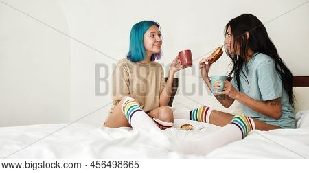 Girl Eating Sweet Bun While Drinking Tea Or Coffee With Her Girlfriend In Bed. Young Lesbian Caucasi