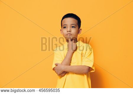 A Little Boy Of Preschool Age Is Standing On An Orange Background In A Yellow Shirt, Looking Thought