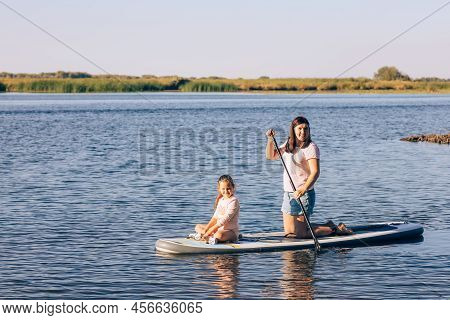 Young Daughter And Mother Sitting Together On Sup Board Rowing With Paddle On Little Lake With Green