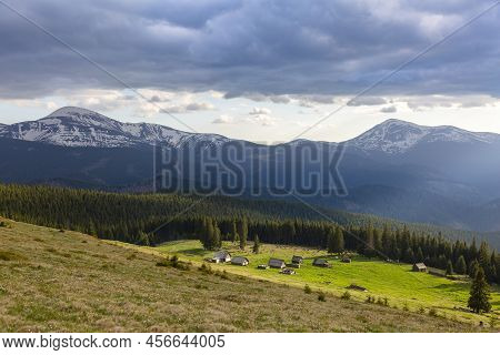 Kukul Green Meadow With Wooden Houses And Forest In Front Of Snowy Peaks, The Carpathians Mountains,