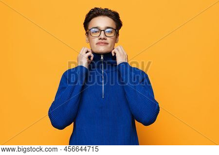 An Attractive Young Man Stands On An Orange Background In A Blue Zip-up Jacket And Black Eyeglasses 