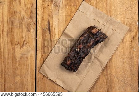Dried Chew Treats For Dogs. Dried Beef Tail Against Wooden Background. Piece Of Tail And Paper Bag F