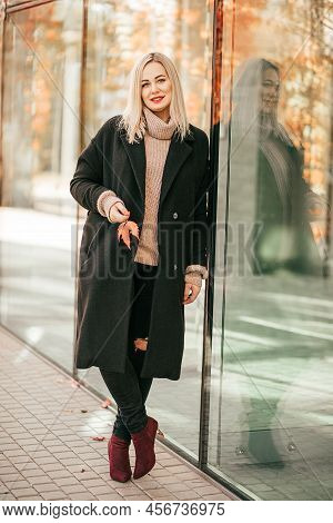 Elegant, Happy Blonde In Black Coat Stands Relaxed Near Glass Windows On Street Of City And Holds Br