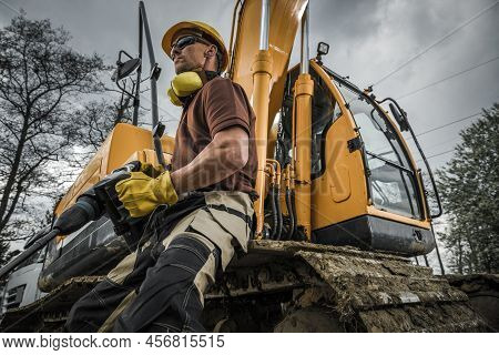 Caucasian Construction Worker In His 40s Proudly Holding A Jackhammer In Front Of Big Excavator Digg
