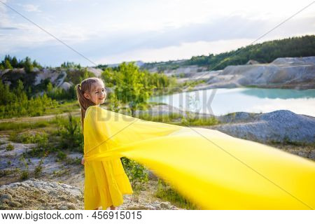 A Small 6-7 Years Old Ukrainian Girl In A Yellow Dress With Wings In Yellow Fabric By The Lake. Blue