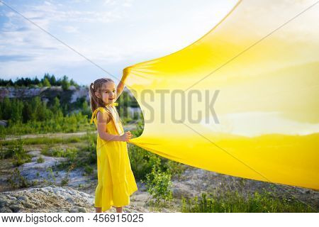 A Small 6-7 Years Old Ukrainian Girl In A Yellow Dress With Wings In Yellow Fabric By The Lake. Blue