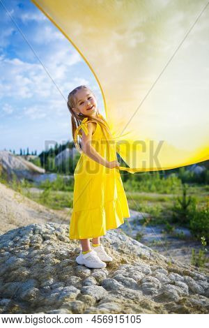 A Small 6-7 Years Old Ukrainian Girl In A Yellow Dress With Wings In Yellow Fabric By The Lake. Blue