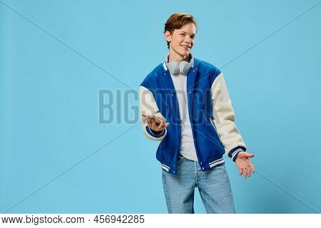 A Stylish Young Man In A Blue Jacket Stands With A Phone In His Hands And Headphones Around His Neck