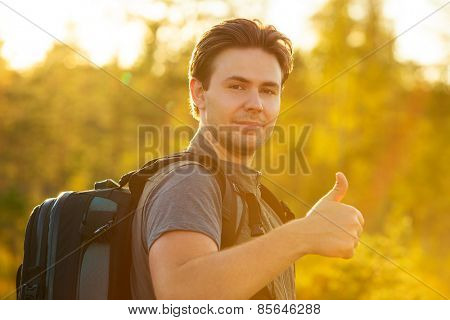 Young man tourist with backpack showing thumbs up handsign. Red sunset light.
