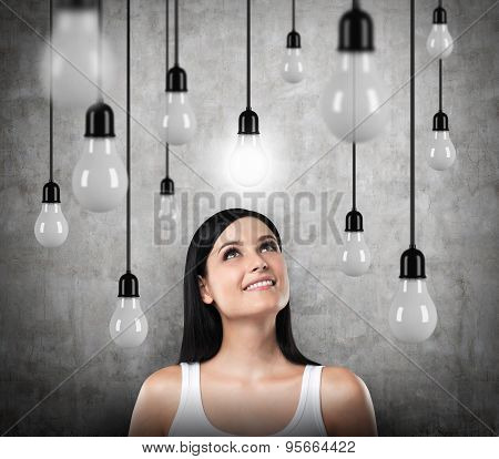 A Dreamy Brunette Looks Upward. Lots Of Light Bulbs, One Of Them Is Turned On. Concrete Background.