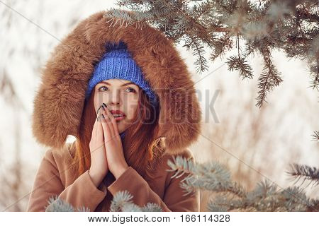 Cute teen girl in blue knitted hat and scarf fur hooded jacket winter walks in the park. She warms the breath of a person hands. Outdoor Activities. Youth fashion. Close-up portrait