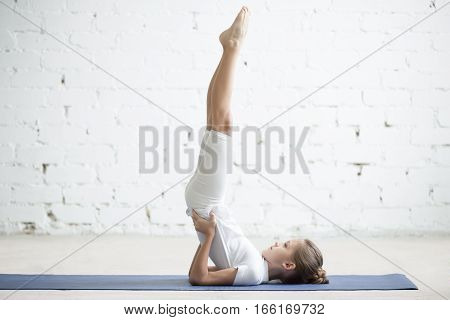 Girl child practicing yoga, standing in salamba sarvangasana exercise, supported Shoulder stand pose, working out wearing sportswear, t-shirt, pants, indoor full length, white loft studio background