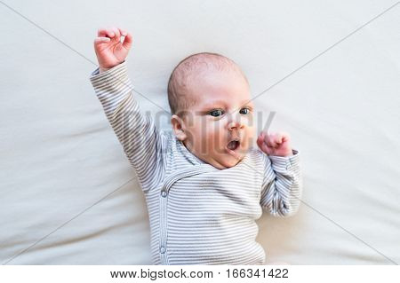 Cute newborn baby boy in striped onesie lying on bed, arm raised, superhero pose, mouth open.