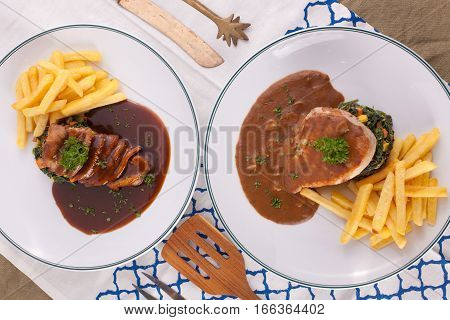 A nice dish of duck confit steak and pork steak served with fried potato, saute spinach and sauce on background with wooden utensils.