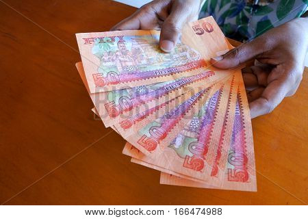 Fijian person counts 50 Fijian dollar bank notes over a wooden counter.
