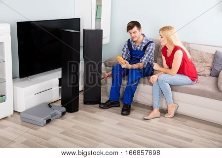 Technician Checking TV Speaker Sound System With Multimeter In Front Of Woman At Home