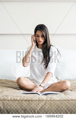 Beautiful woman in a shirt talking on the phone and reading the morning paper on the luxorious bedroom bed