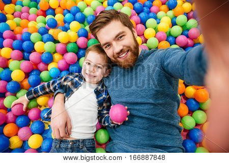 Happy little son with his dad having fun and taking selfie lying in pool with colorful balls