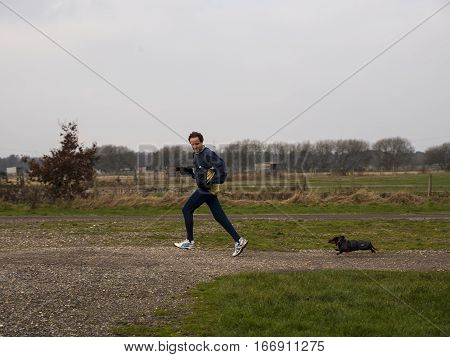 Man in winter running gear running off road looking over his shoulder where he is followed by miniature dachshund