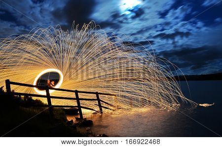 A long exposure of steel wool spinning with sparks by a fence and reflected in a lake