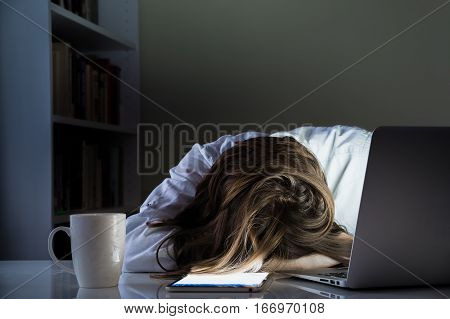 Tired person sleeping at desk in home office with turned on computer and tablet PC. Exhausted female person taking nap in dark room of home office overworking with laptop