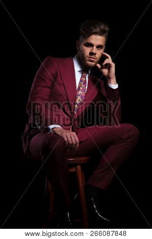 pensive young elegant man in burgundy suit and tie sitting on a stool