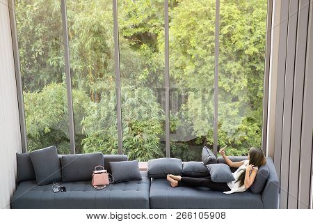 Asian Woman Laying On Sofa Near Big Glass Wondows, Relaxing Alone In House With Green Forest In Back