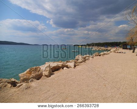 Stone Beach In Sibenik On A Summer Day. Rocks And Two Boats Are Visible. In The Backbround Are Sunnb