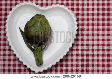 Purple Fresh Artichoke On A Heart Shape White Plate On  Red And White Chekered Tablecloth