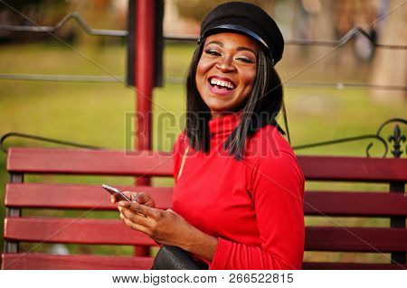 African American Fashion Girl In Newsboy Cap, With Handbag Posed At Street, Sitting On Bench With Mo