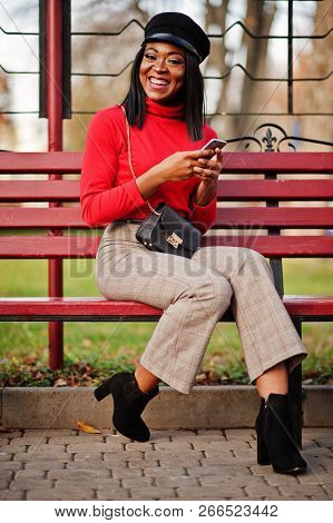African American Fashion Girl In Newsboy Cap, With Handbag Posed At Street, Sitting On Bench With Mo