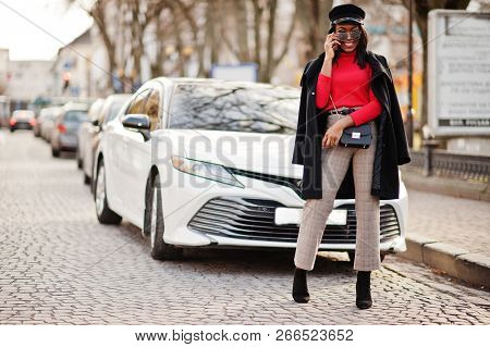 African American Fashion Girl In Coat, Newsboy Cap And Sunglasses Posed At Street Against White Busi