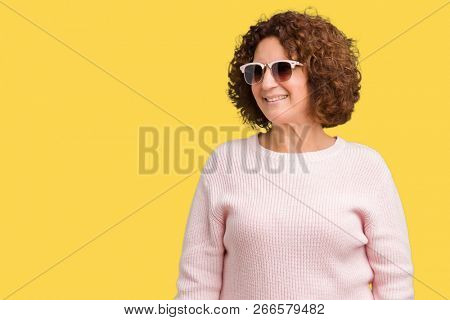 Beautiful middle ager senior woman wearing pink sweater and sunglasses over isolated background looking away to side with smile on face, natural expression. Laughing confident.