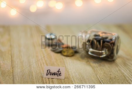 Donate Money Concept With Shiny Coins On A Glass Jar, Soft Bokeh Background On Wooden Board