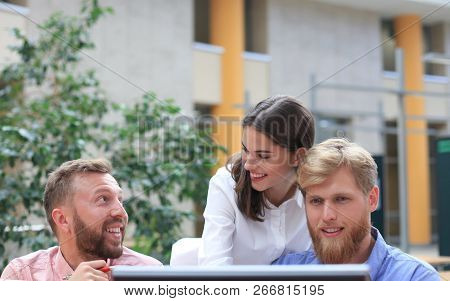 Group Of Young People In Casual Wear Sitting At The Office Desk And Discussing Something While Looki