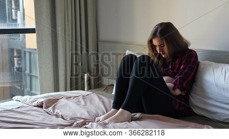 Young Woman Sits On Large Bed Writhes Holding Hands On Stomach In Bedroom
