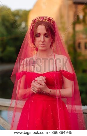 Portrait Of Attractive Redhead Tattooed Woman In Red Dress, Diadema And Red Veil On Blurred Medieval