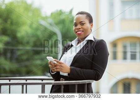 Using Smartphone. African-american Businesswoman In Office Attire Smiling, Looks Confident And Happy