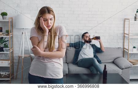 Alcoholism And Tyranny In Family. Unhappy Woman Presses Her Hand To Cheek, Man Sits On Sofa, Watches