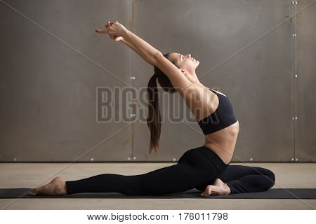 Young attractive woman practicing yoga, stretching in One Legged King Pigeon exercise, Eka Pada Raja Kapotasana pose, working out, wearing black sportswear, cool urban style, full length, grey studio