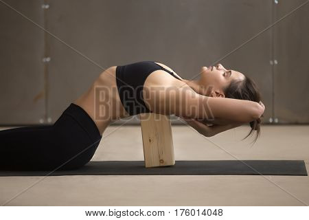 Young attractive sporty woman practicing yoga, stretching in Matsyasana using wooden block, Fish pose, exercise for spine and shoulders flexibility working out, cool urban style, grey studio, closeup