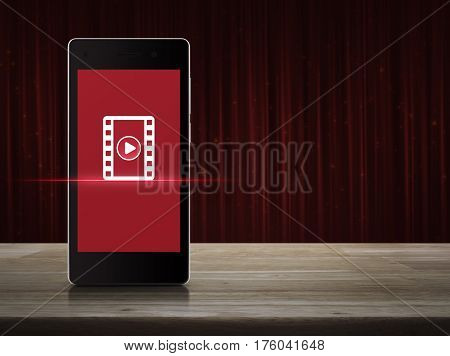 Play button with movie icon on modern smart phone screen on wooden table over red curtain with sparkle glitter Cinema online concept
