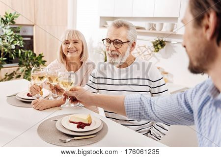 Happy together. Charismatic optimistic cheerful aged couple having dinner and enjoying weekend with their mature son while holding glasses full of champagne