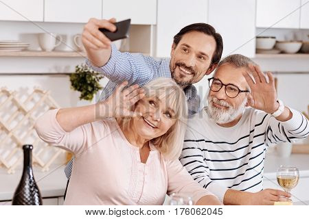 Full of joy. Happy young smiling man having dinner and enjoying free time with his aged parents while holding the mobile and making selfie