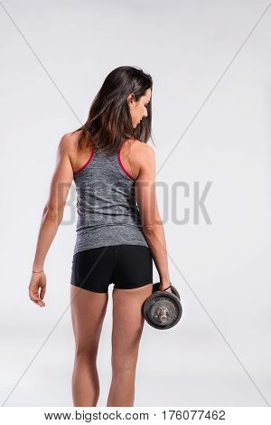 Attractive young fitness woman in tank top and black shorts, holding dumbbell. Slim waist, perfect fit female body. Rear view. Studio shot on gray background.