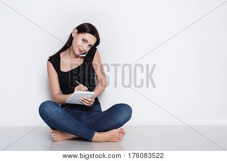 Beautiful business woman in casual clothes talking on mobile phone and taking notes while sitting on floor at home and working. Girl making call to delivery, white studio background, copy space