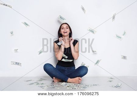 Excited woman sitting under money rain sit on floor and smile. Happy girl drop american dollar banknotes in the air - lottery jackpot and financial success concept, white studio background
